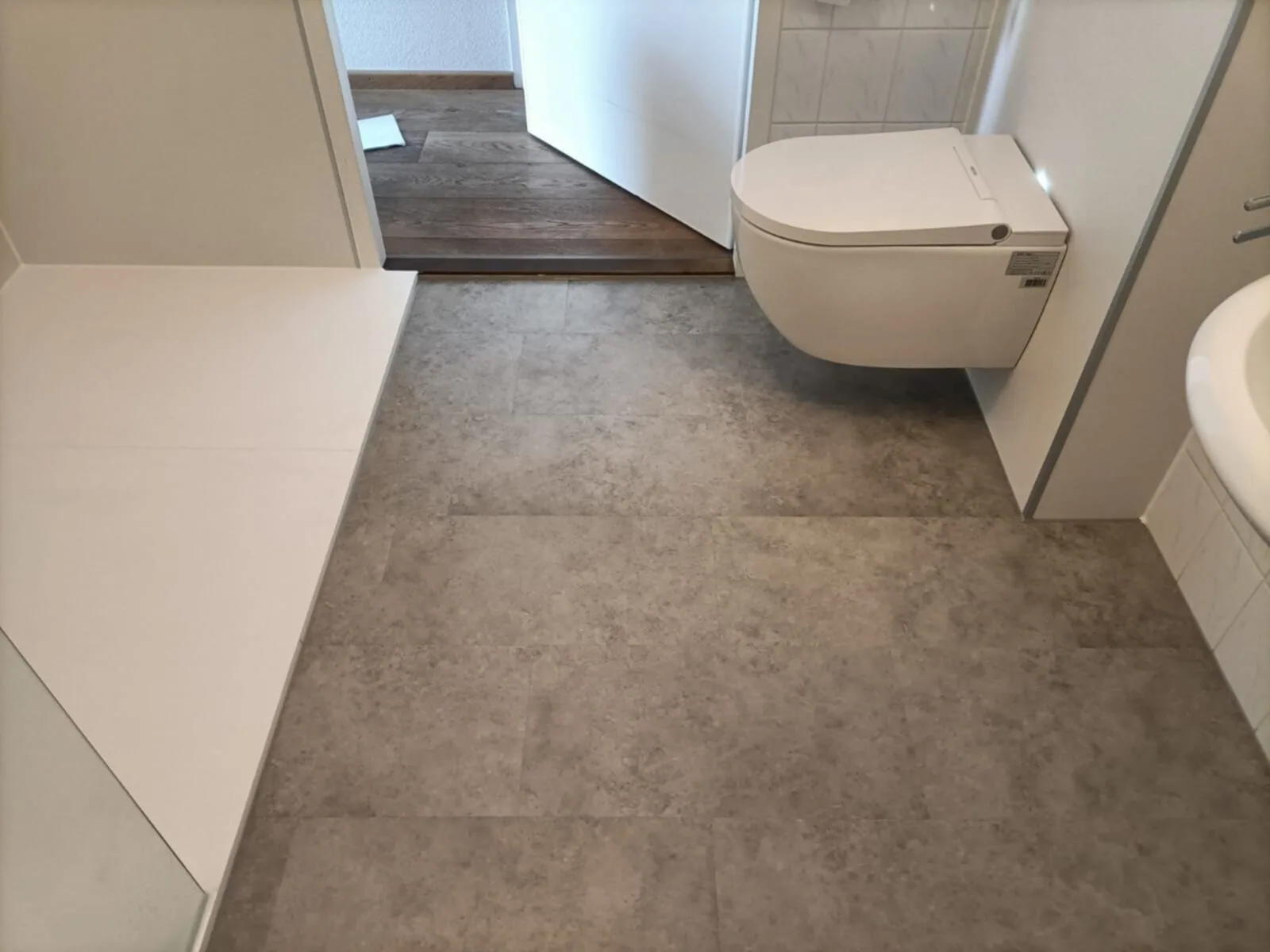 Wandhangend douchetoilet naast een witte douchevloer op een grijze steenlook vloer