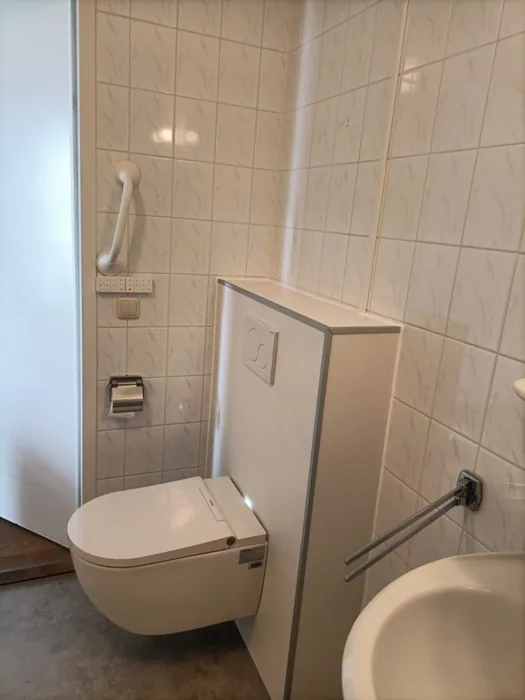 Wandhangend douchetoilet tegen een witte voorzetwand met steunbeugel en tegels