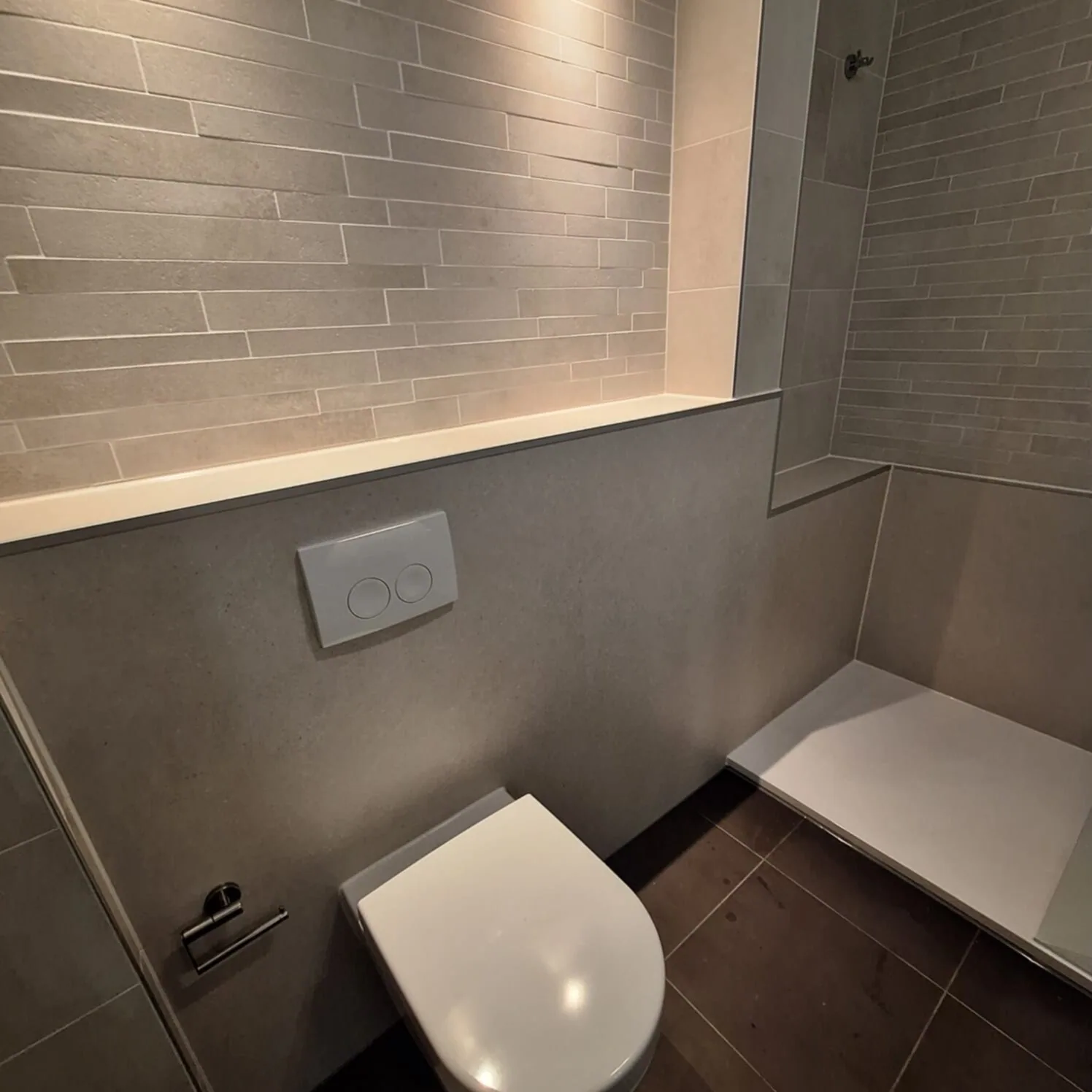 Hangtoilet tegen een grijze voorzetwand met verlichte nis, rechts een witte douchevloer