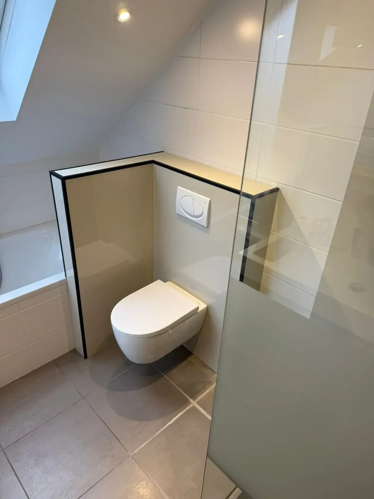 Hangtoilet op een voorzetwand met zwarte afwerkrand onder een schuin dak, naast ligbad en glazen wand