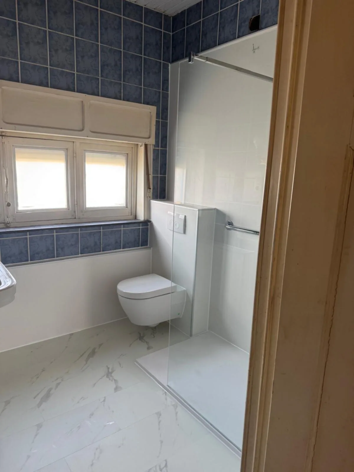Inloopdouche met glazen wand, hangtoilet met voorzetwand en steunbeugel, marmerlook vloer en blauwe tegelband