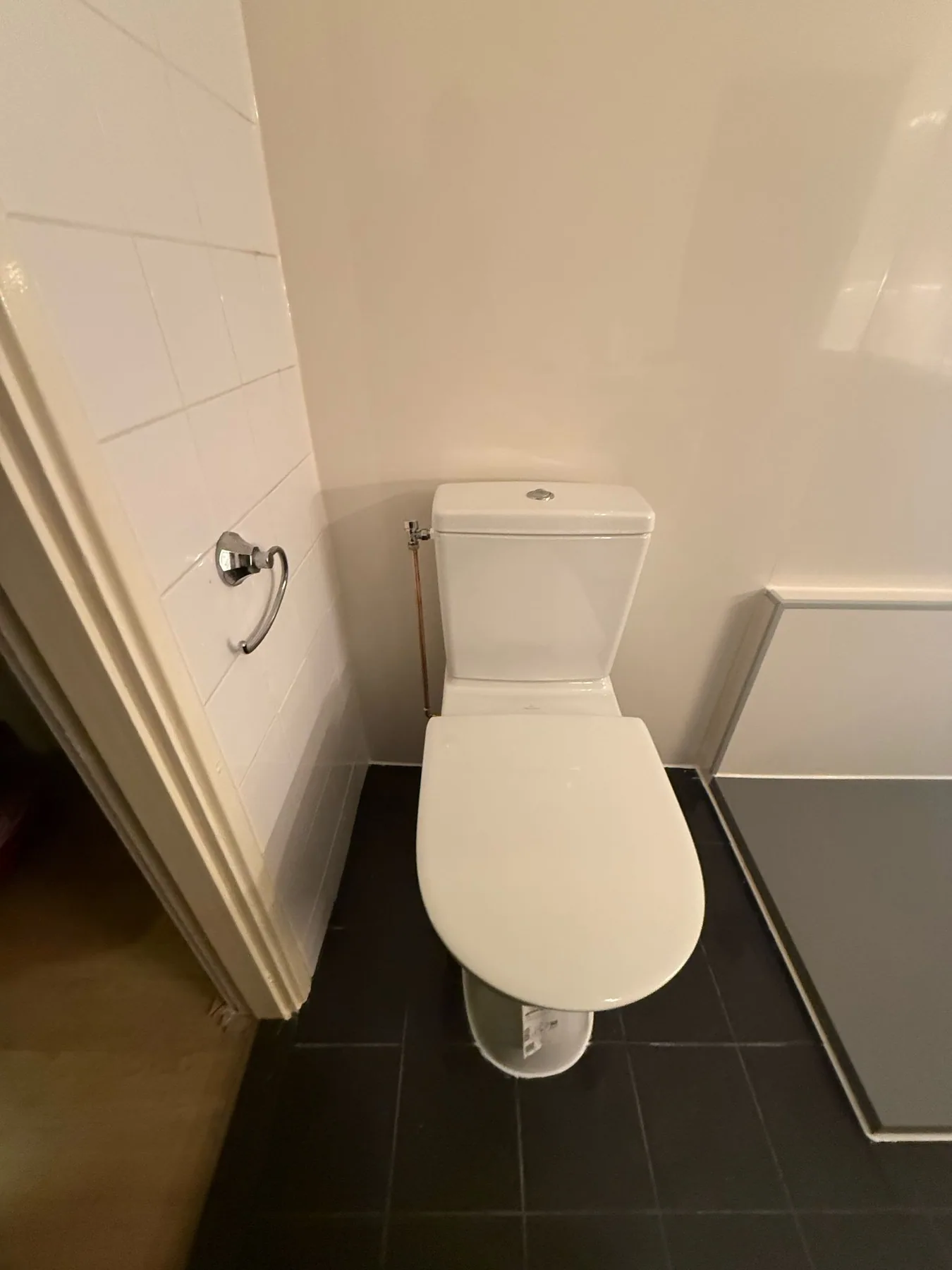Wit staand toilet op donkere vloertegels naast een witte wand en deurkozijn