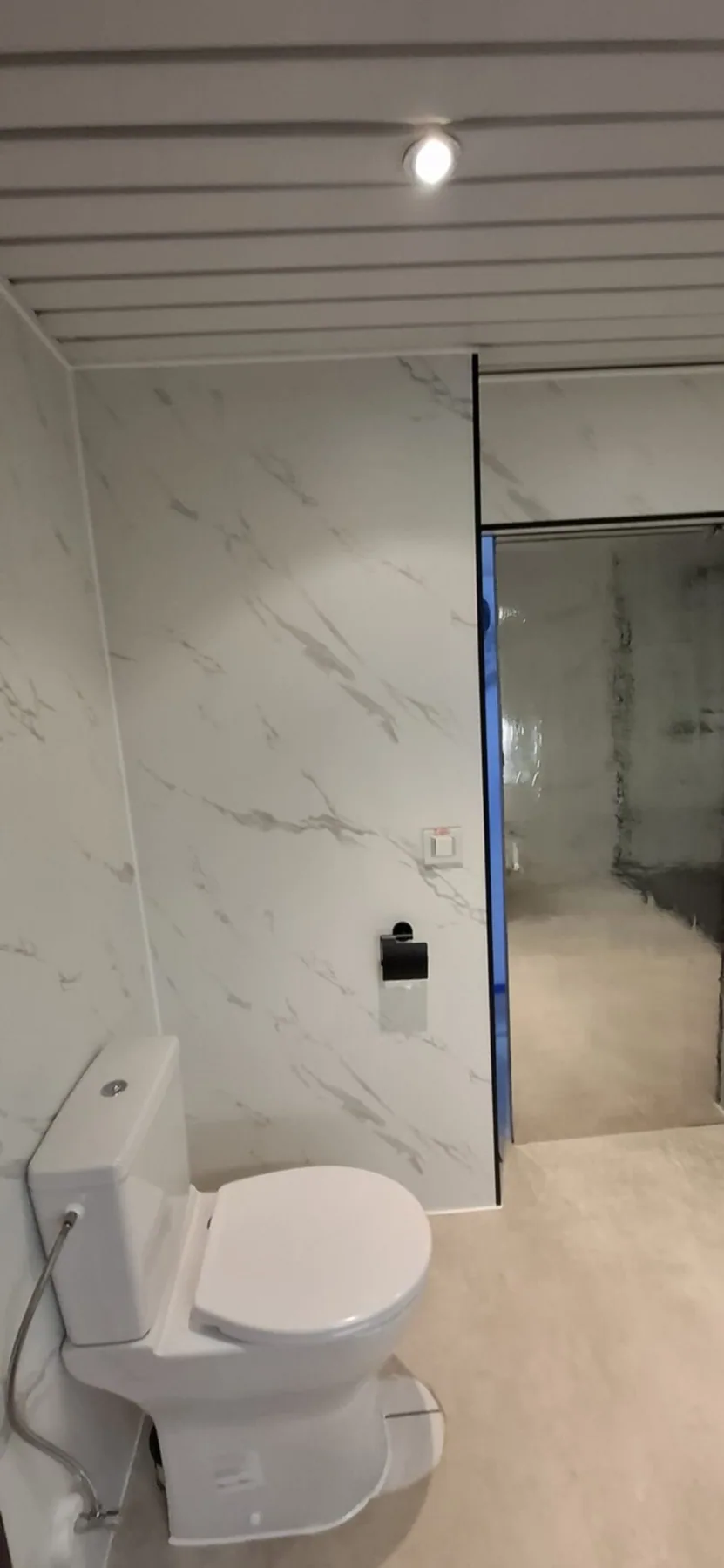 Staand toilet met zwarte rolhouder tegen witte marmerlook wandpanelen, lamellenplafond met spot