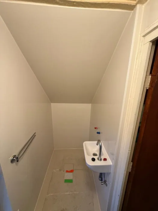 Smalle toiletruimte onder de trap met nieuwe witte wanden, fonteintje en beugel, het toilet nog niet geplaatst