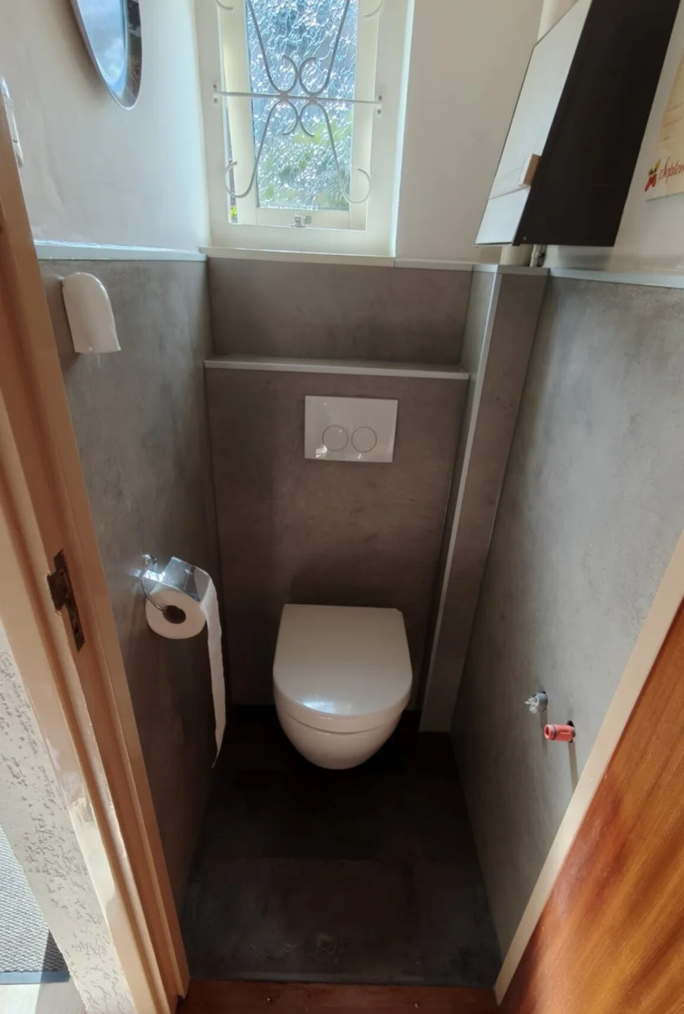 Dezelfde toiletruimte na de renovatie: hangtoilet, voorzetwand met drukplaat en grijze betonlook wandpanelen