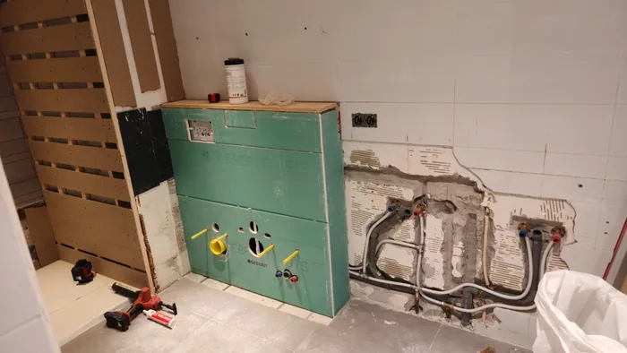 Groene voorzetwand in aanbouw met twee uitsparingen voor toilet en bidet, oude leidingen ernaast
